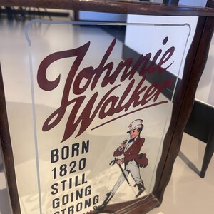 K&ouml;nnte beinhalten: Ein Vintage Johnnie Walker Spiegel-Schild mit einem dunklen Holzrahmen. Das Schild zeigt den Markennamen in burgunderfarbener Schrift, den Text "BORN 1820 STILL GOING STRONG" und eine Illustration eines Mannes in rotem Mantel und Zylinder.