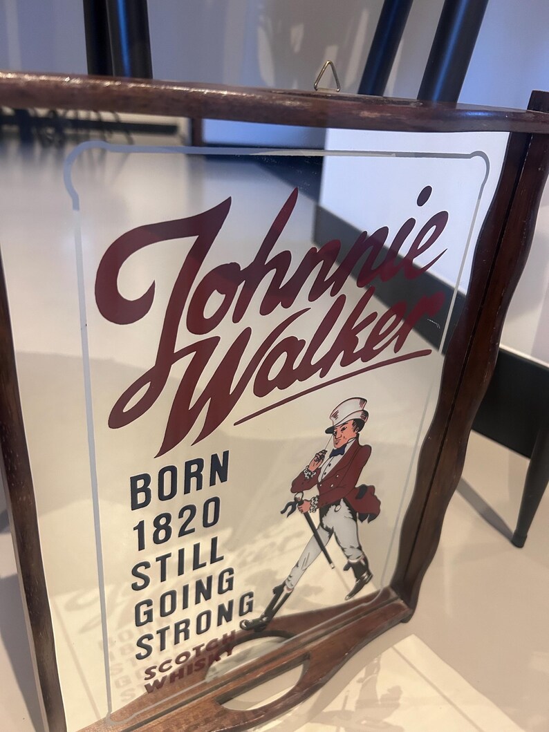 K&ouml;nnte beinhalten: Ein Vintage Johnnie Walker Spiegel-Schild mit einem dunklen Holzrahmen. Das Schild zeigt den Markennamen in burgunderfarbener Schrift, den Text "BORN 1820 STILL GOING STRONG" und eine Illustration eines Mannes in rotem Mantel und Zylinder.