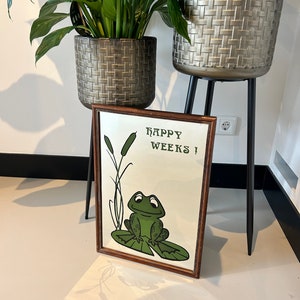 May include: A framed mirror with a brown wooden frame. The mirror has a green frog sitting on a lily pad with the text "Happy Weeks!" printed on the mirror.
