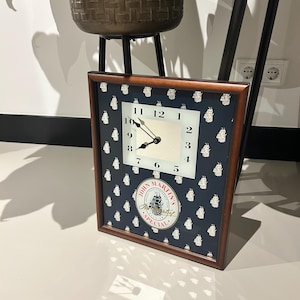 Op de afbeelding: Een bruine ingelijste klok met een witte wijzerplaat en zwarte wijzers. De klok heeft een donkerblauwe achtergrond met een wit patroon van schepen. De wijzerplaat van de klok heeft de tekst "John Martin's Special" in een cirkel.