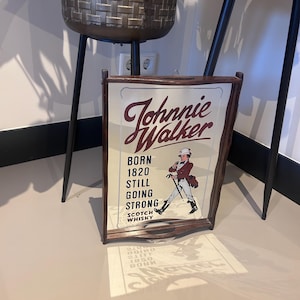 K&ouml;nnte beinhalten: Holzgerahmtes Schild mit dem Johnnie Walker-Logo und dem Text "Born 1820 Still Going Strong". Das Schild zeigt eine Illustration eines Mannes in einem roten Mantel und Zylinder. Das Schild lehnt an einer Wand.