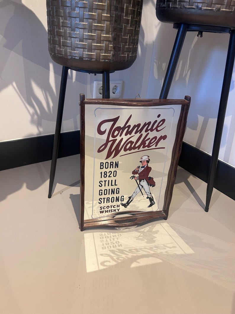 K&ouml;nnte beinhalten: Holzgerahmtes Schild mit dem Johnnie Walker-Logo und dem Text "Born 1820 Still Going Strong". Das Schild zeigt eine Illustration eines Mannes in einem roten Mantel und Zylinder. Das Schild lehnt an einer Wand.