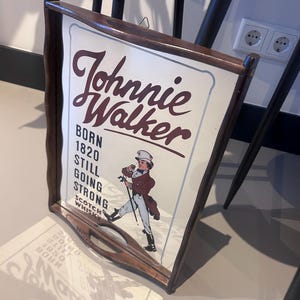 K&ouml;nnte beinhalten: Ein Vintage-Schild f&uuml;r Johnnie Walker Scotch Whisky. Das Schild zeigt den Markennamen in einer stilisierten Schriftart zusammen mit dem Text "BORN 1820 STILL GOING STRONG". Es enth&auml;lt eine Illustration eines Mannes mit Zylinder und Mantel, der mit einem Stock geht. Das Schild ist in dunklem Holz gerahmt.