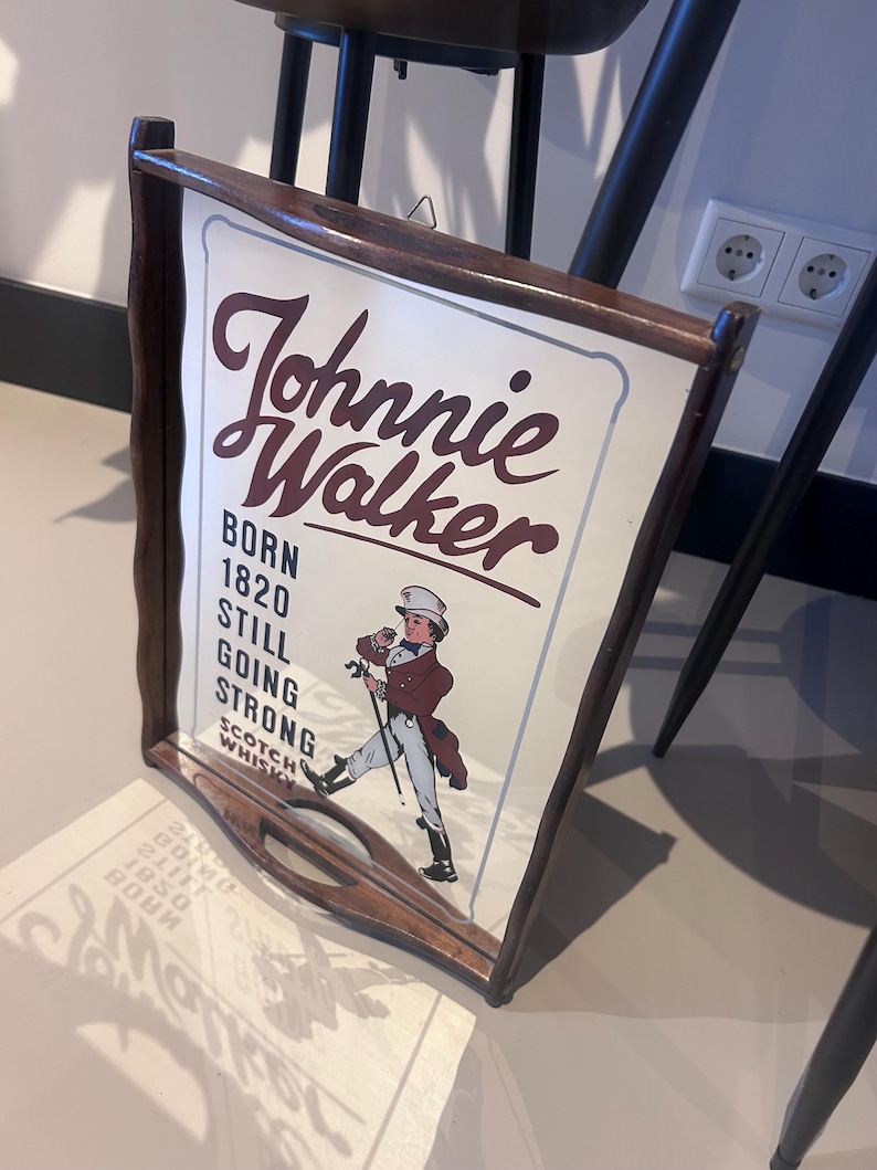 K&ouml;nnte beinhalten: Ein Vintage-Schild f&uuml;r Johnnie Walker Scotch Whisky. Das Schild zeigt den Markennamen in einer stilisierten Schriftart zusammen mit dem Text "BORN 1820 STILL GOING STRONG". Es enth&auml;lt eine Illustration eines Mannes mit Zylinder und Mantel, der mit einem Stock geht. Das Schild ist in dunklem Holz gerahmt.