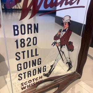 K&ouml;nnte beinhalten: Vintage John Walker Scotch Whisky Werbeschild. Das Schild zeigt die Worte "Walker" in roter Schrift und den Text "BORN 1820 STILL GOING STRONG SCOTCH WHISKY". Es enth&auml;lt auch eine Illustration eines Mannes in einem roten Mantel und Zylinder.