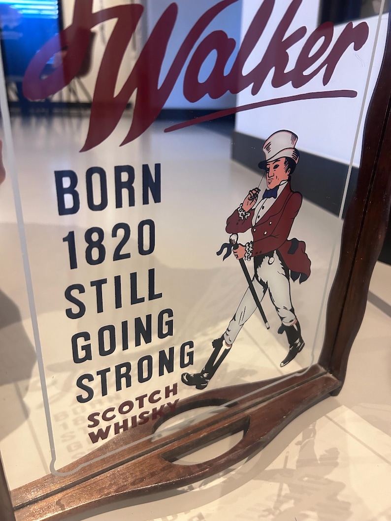 K&ouml;nnte beinhalten: Vintage John Walker Scotch Whisky Werbeschild. Das Schild zeigt die Worte "Walker" in roter Schrift und den Text "BORN 1820 STILL GOING STRONG SCOTCH WHISKY". Es enth&auml;lt auch eine Illustration eines Mannes in einem roten Mantel und Zylinder.