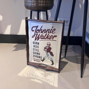 K&ouml;nnte beinhalten: Ein gerahmtes Schild f&uuml;r Johnnie Walker Scotch Whisky. Das Schild zeigt das Logo der Marke und eine Darstellung eines Mannes in einem roten Mantel und Zylinder, der mit einem Stock geht. Der Text auf dem Schild lautet "Born 1820, Still Going Strong."