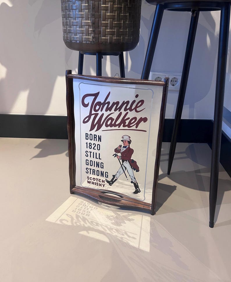 K&ouml;nnte beinhalten: Ein gerahmtes Schild f&uuml;r Johnnie Walker Scotch Whisky. Das Schild zeigt das Logo der Marke und eine Darstellung eines Mannes in einem roten Mantel und Zylinder, der mit einem Stock geht. Der Text auf dem Schild lautet "Born 1820, Still Going Strong."