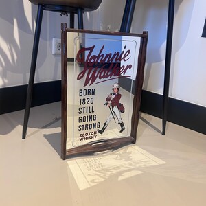 K&ouml;nnte beinhalten: Gerahmter Spiegel mit dem Johnnie Walker Logo und Text. Der Spiegel hat einen dunkelbraunen Holzrahmen. Der Text lautet "BORN 1820 STILL GOING STRONG SCOTCH WHISKY" und eine Illustration eines Mannes in einem roten Mantel.