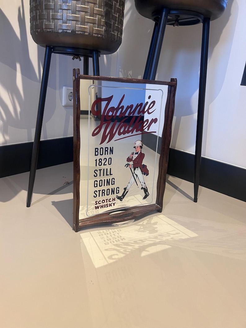 K&ouml;nnte beinhalten: Gerahmter Spiegel mit dem Johnnie Walker Logo und Text. Der Spiegel hat einen dunkelbraunen Holzrahmen. Der Text lautet "BORN 1820 STILL GOING STRONG SCOTCH WHISKY" und eine Illustration eines Mannes in einem roten Mantel.