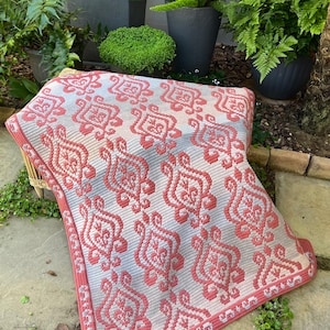 May include: A crocheted blanket featuring a repeating coral floral design on a light grey backdrop. The rectangular blanket has a coral border. It is draped over a wicker chair, with plants in the background. The blanket measures approximately 150cm x 100cm.