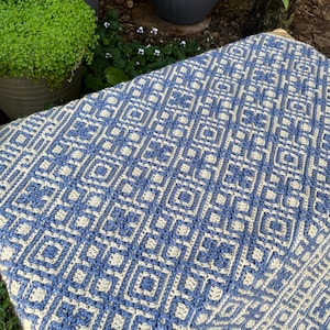 May include: A blue and cream crocheted blanket with a geometric diamond pattern. The blanket is folded over a surface, displaying the intricate design and texture. The colour palette is a mix of light blue and cream, creating a visually appealing textile.