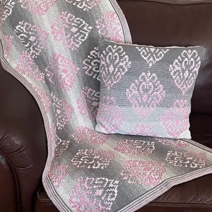 May include: A gray and pink crocheted throw blanket with a matching pillow. The blanket and pillow feature a repeating pattern of stylized flowers.