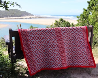 May include: A red and white crocheted throw blanket with a geometric pattern is draped over a wooden bench. The bench is positioned outdoors with a view of a sandy beach and ocean.