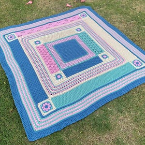 May include: A crocheted blanket with a blue, pink, and white color scheme. The blanket is made up of squares with different patterns and textures.