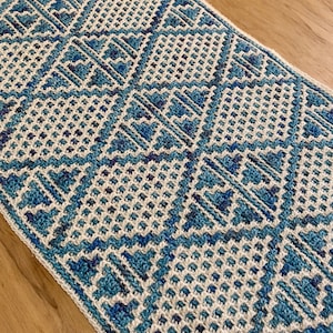 Pode incluir: Uma manta de crochê branca e azul com um padrão geométrico. A manta é feita com uma variedade de pontos e tem uma superfície texturizada.