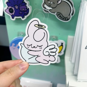 May include: A light pink sticker featuring a cartoon angel bunny with closed eyes, a halo, and wings. The sticker is held by a hand, with other stickers visible in the background.