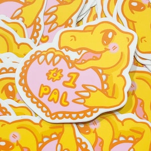 May include: A yellow cartoon dinosaur sticker holding a pink heart with the text "# 1 PAL" in orange. The dinosaur has a friendly expression with rosy cheeks. The sticker has a white border and is surrounded by other similar stickers.