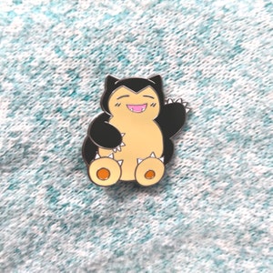 May include: A Snorlax enamel pin with a black and tan color scheme. The pin features a smiling Snorlax with its arm raised in a wave. The pin is set against a light blue and white textured background.