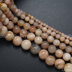 May include: A strand of sunstone beads in various sizes. The beads are round and have a light peach color with white streaks. The beads are arranged in a line, with the largest beads at the top and the smallest beads at the bottom.