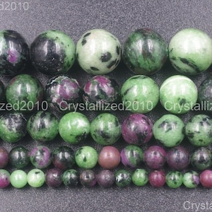 May include: A strand of green and purple gemstone beads. The beads are round and have a speckled pattern. The beads are arranged in a row and are separated by a small space. The beads are all the same size and shape. The beads are a mix of green and purple colours. The beads are made of natural stone.