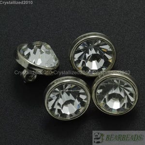 Puede incluir: Cuatro botones redondos plateados con centros de cristal transparente. Los botones tienen un borde elevado alrededor del cristal.