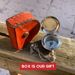 Peut inclure: Une boussole argentée avec un cadran blanc et des marques noires. La boussole est attachée à une chaîne argentée et se trouve dans un petit étui en cuir orange. L'étui a un rabat qui se ferme à l'aide d'un bouton. Le texte "BOX IS OUR GIFT" est imprimé sur une bannière rouge.