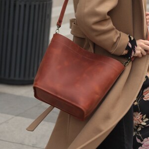 May include: A woman wearing a camel-colored coat with a floral lining and black pants. She is carrying a brown leather bucket bag with a long strap.