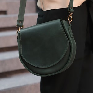 K&ouml;nnte beinhalten: Eine dunkelgr&uuml;ne Ledertasche mit einem langen Riemen. Die Tasche hat einen Klappverschluss und eine goldene Schnalle.