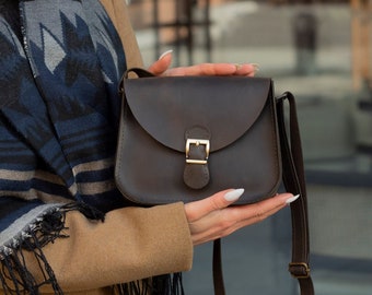 May include: A woman is holding a brown leather crossbody bag with a gold buckle. The bag has a long strap and is closed with a flap.