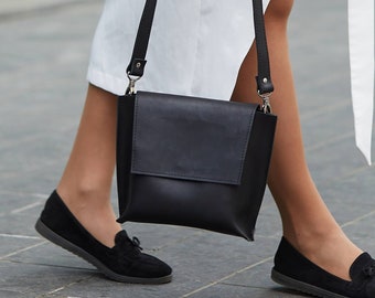 Può includere: Una donna con un vestito bianco e mocassini neri porta una borsa a tracolla in pelle nera con una tracolla.