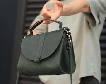 Pode incluir: Uma bolsa de mão de couro verde escuro com alça superior e alça longa. A bolsa tem fecho de aba com fivela.