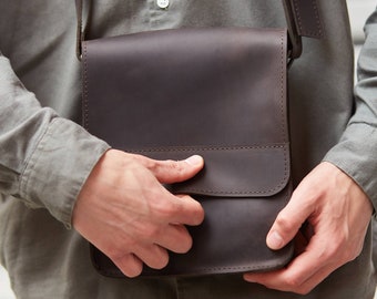 May include: A brown leather messenger bag with a flap closure and a strap. The bag is being held by a person wearing a gray shirt and beige pants.