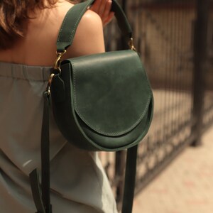 K&ouml;nnte beinhalten: Eine dunkelgr&uuml;ne Lederschultertasche mit einer geschwungenen Klappe und einer goldenen Schnalle. Die Tasche wird von einer Person mit einem hellblauen Oberteil und einer dunklen Hose getragen.