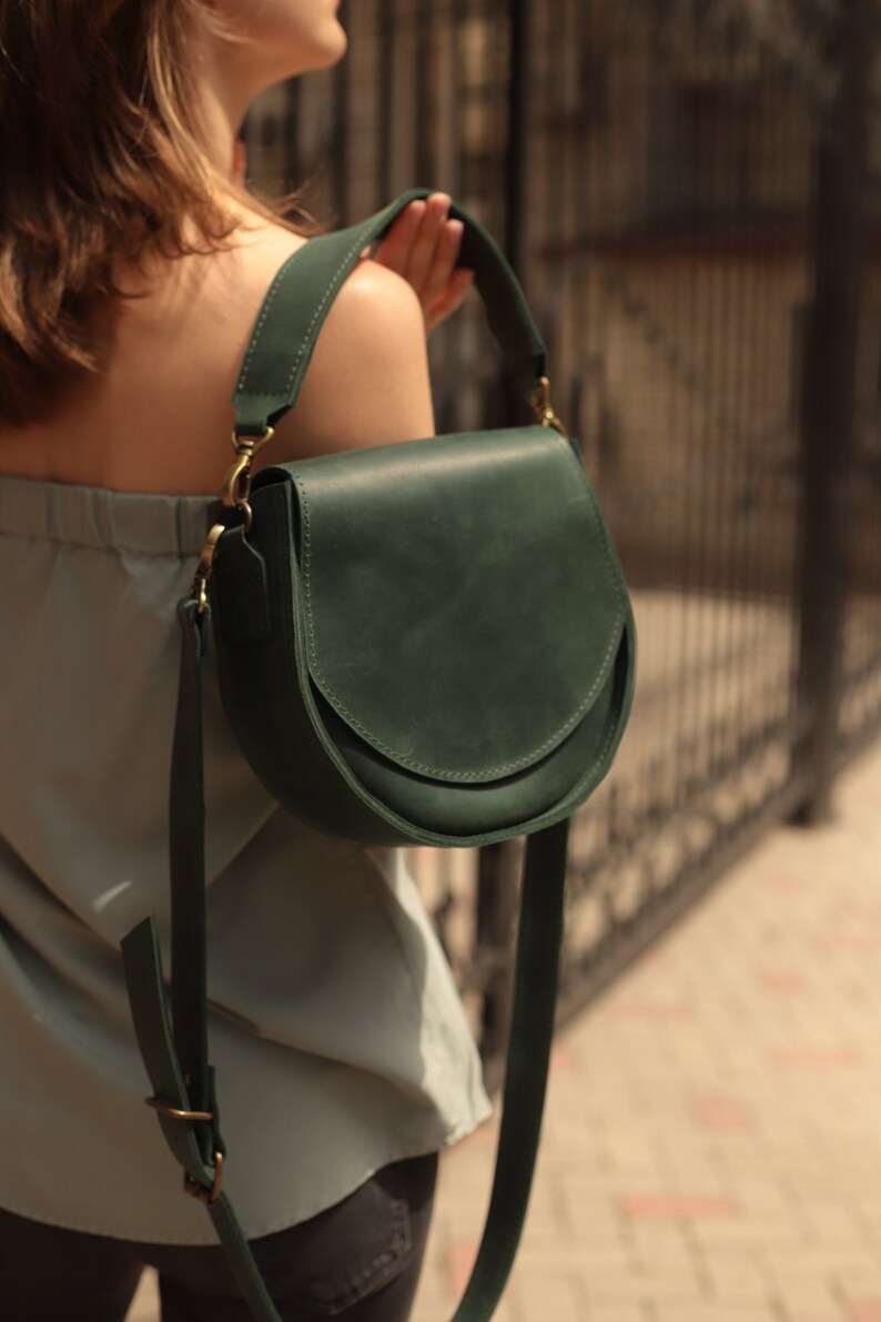 K&ouml;nnte beinhalten: Eine dunkelgr&uuml;ne Lederschultertasche mit einer geschwungenen Klappe und einer goldenen Schnalle. Die Tasche wird von einer Person mit einem hellblauen Oberteil und einer dunklen Hose getragen.