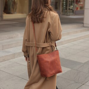May include: A woman wearing a camel-colored coat and black pants walks down the street. She is carrying a brown leather satchel bag with a long strap.