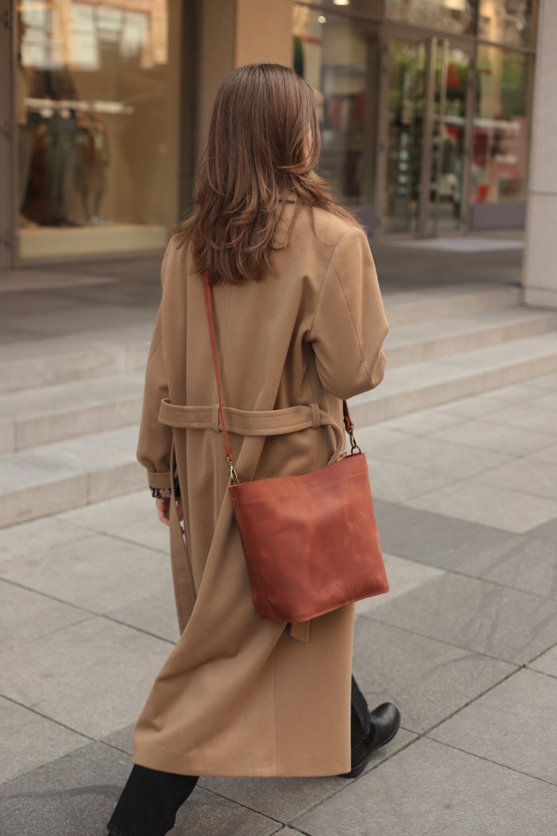 May include: A woman wearing a camel-colored coat and black pants walks down the street. She is carrying a brown leather satchel bag with a long strap.