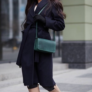 May include: A woman wearing a navy blue coat with a belt, a black beret, and knee-high beige boots. She is carrying a dark green leather crossbody bag.