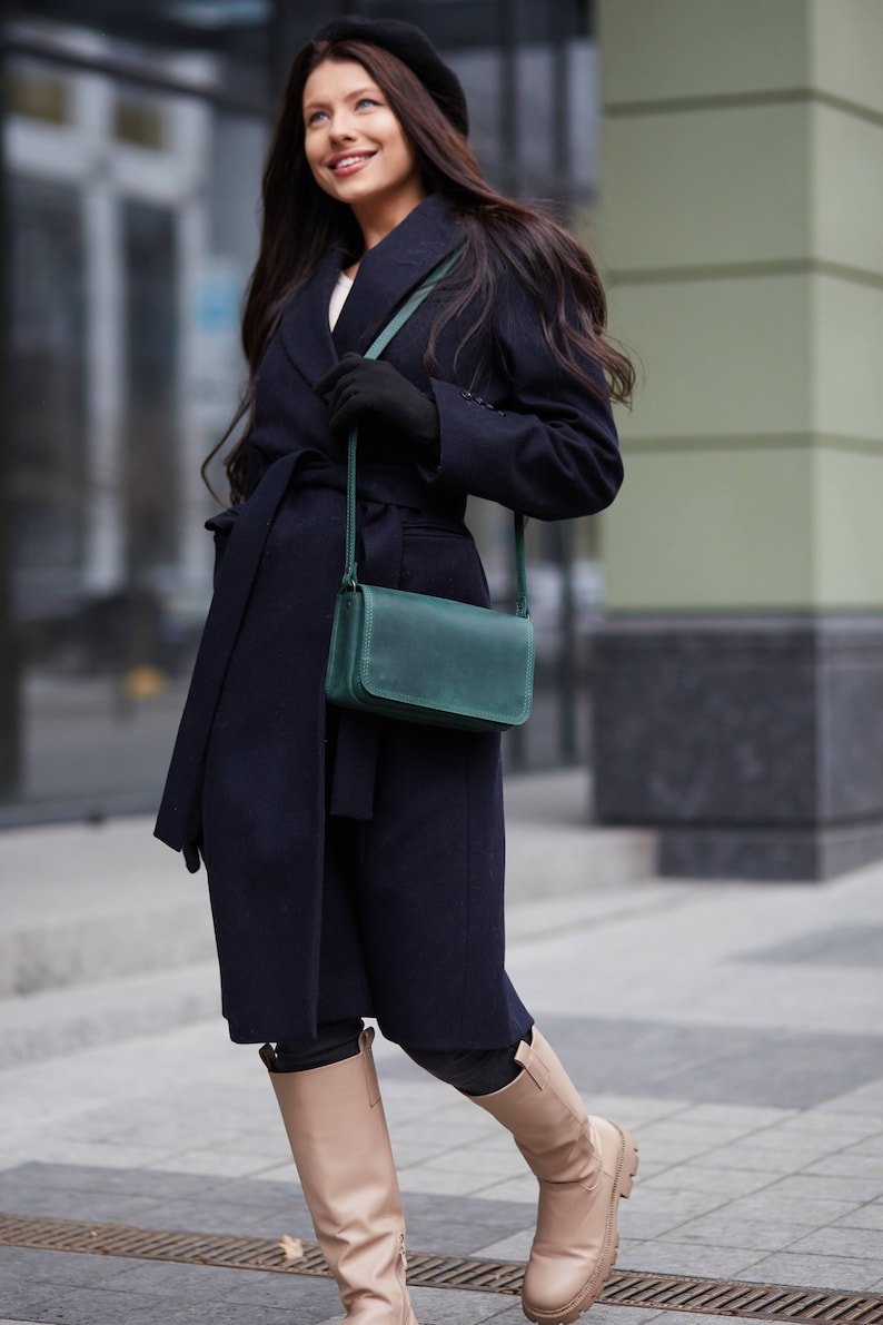 May include: A woman wearing a navy blue coat with a belt, a black beret, and knee-high beige boots. She is carrying a dark green leather crossbody bag.