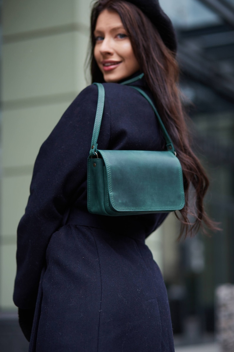 May include: A woman wearing a black coat and a beret carries a dark green leather shoulder bag. The bag has a flap closure and a single strap.