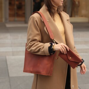May include: A woman wearing a camel-colored coat with a brown leather tote bag and a brown leather wallet.