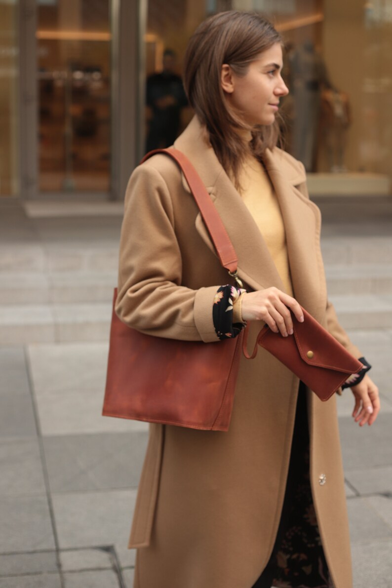 May include: A woman wearing a camel-colored coat with a brown leather tote bag and a brown leather wallet.
