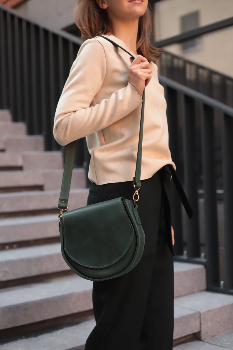 K&ouml;nnte beinhalten: Eine dunkelgr&uuml;ne Ledertasche mit einem langen Riemen. Die Tasche hat einen Klappverschluss und eine goldene Schnalle.