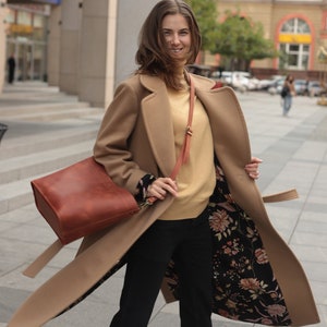 May include: A woman wearing a camel-colored coat with a floral lining. The coat has a belt and a brown leather bag is slung over her shoulder.