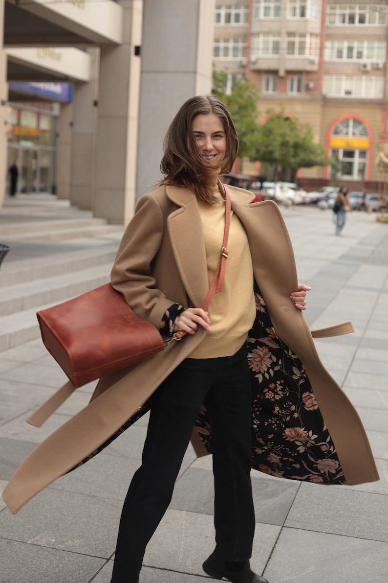 May include: A woman wearing a camel-colored coat with a floral lining. The coat has a belt and a brown leather bag is slung over her shoulder.
