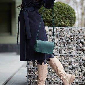 May include: A woman wearing a black coat with a green leather crossbody bag and beige knee-high boots.