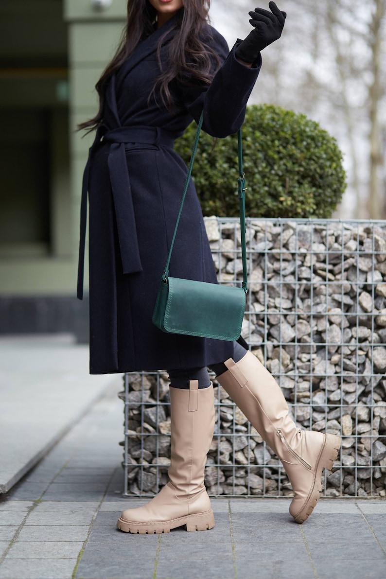 May include: A woman wearing a black coat with a green leather crossbody bag and beige knee-high boots.