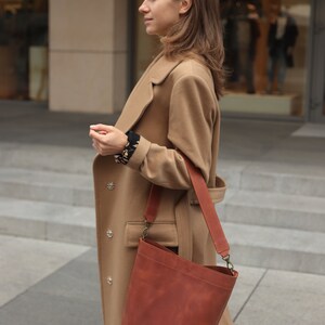 May include: A woman wearing a camel-colored coat with a brown leather bucket bag. The bag has a long strap and a buckle closure.