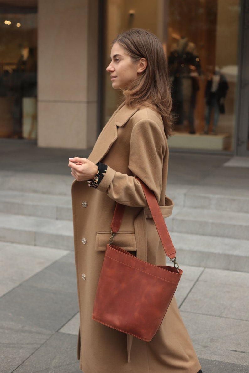 May include: A woman wearing a camel-colored coat with a brown leather bucket bag. The bag has a long strap and a buckle closure.