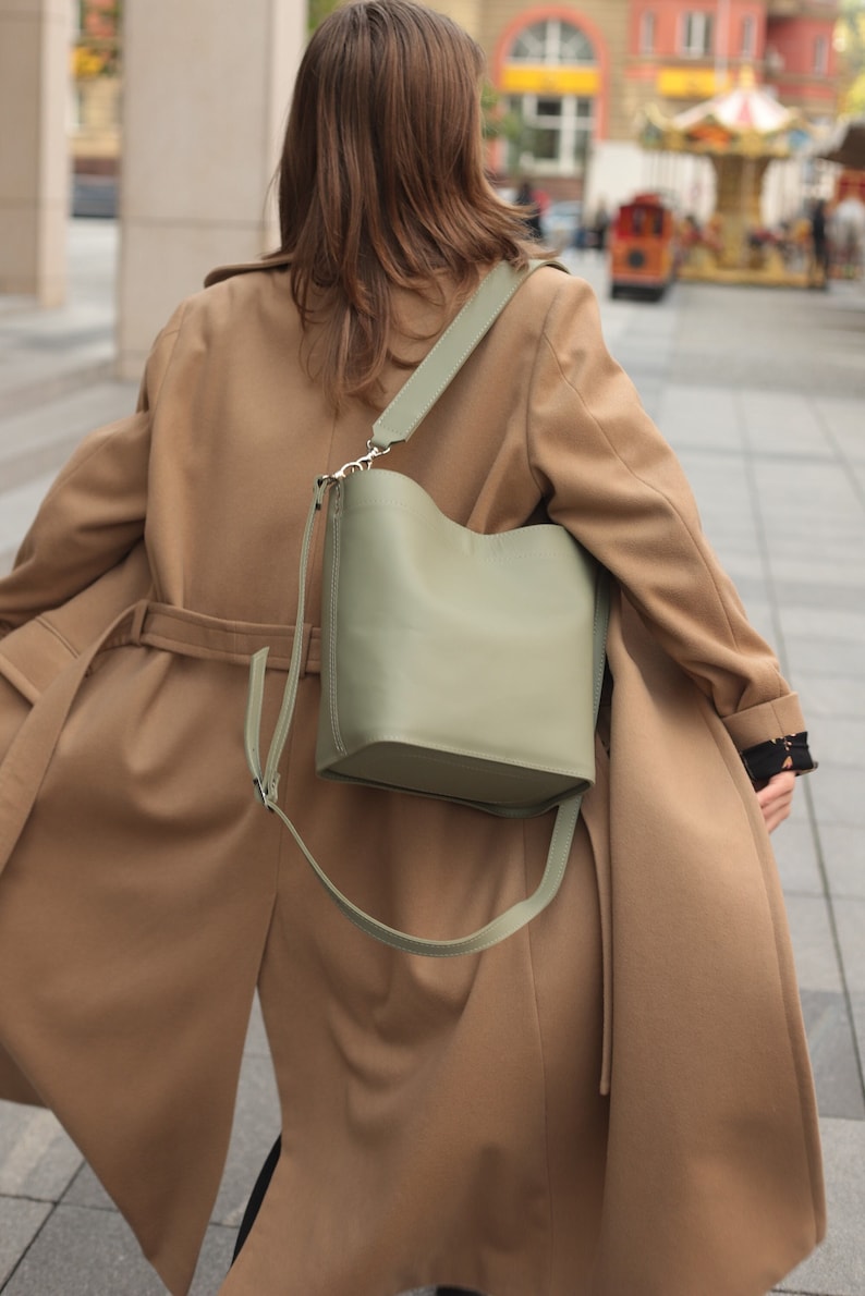 May include: A woman wearing a brown coat with a green leather bucket bag slung over her shoulder.